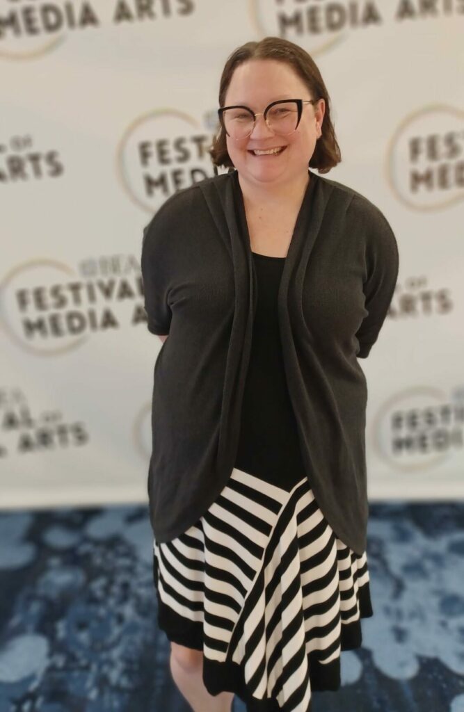 A woman with short brown hair and glasses smiles in front of the BEA Festival of Media repeatable